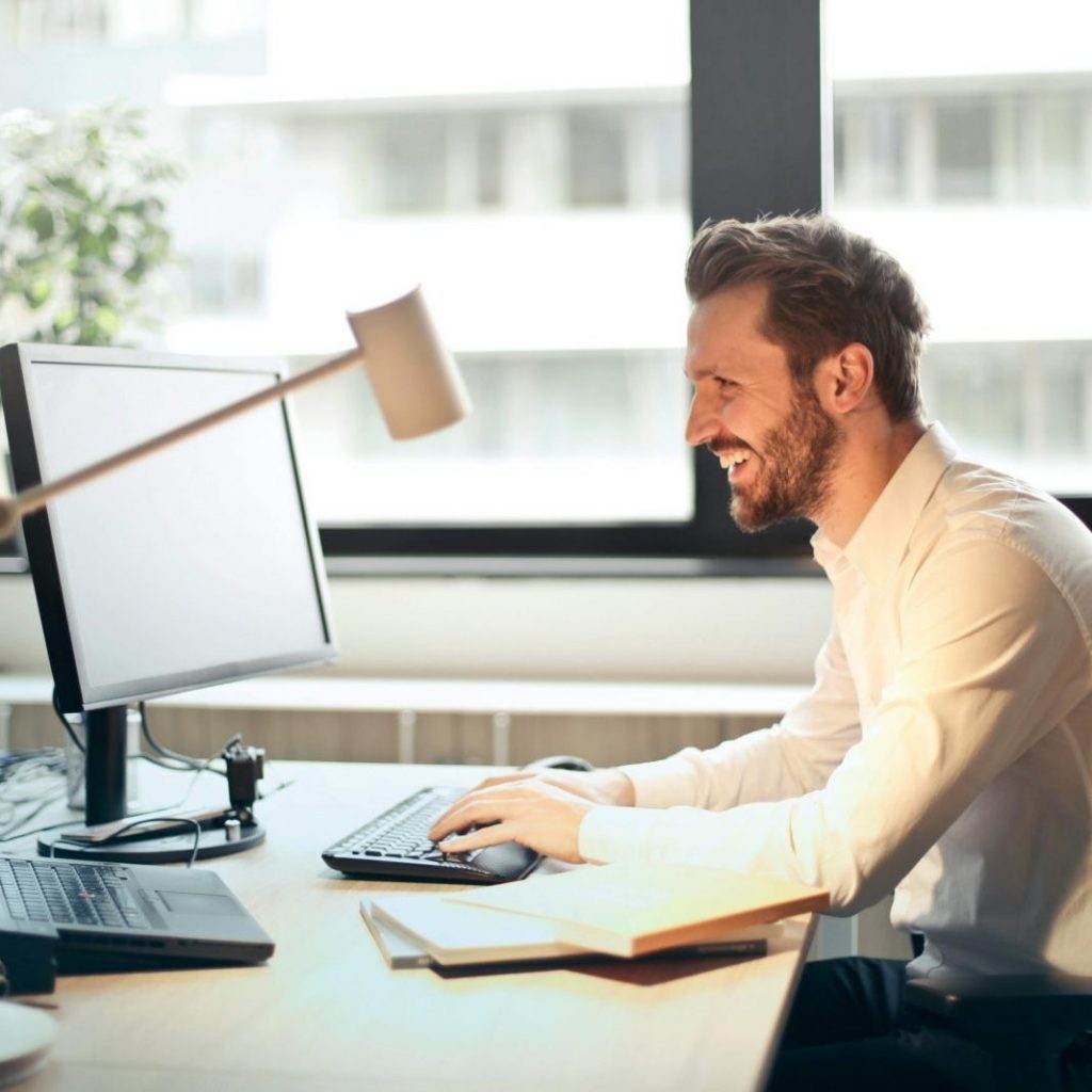 A man smiling while working at an office desk with a computer and natural daylight streaming in through large windows.